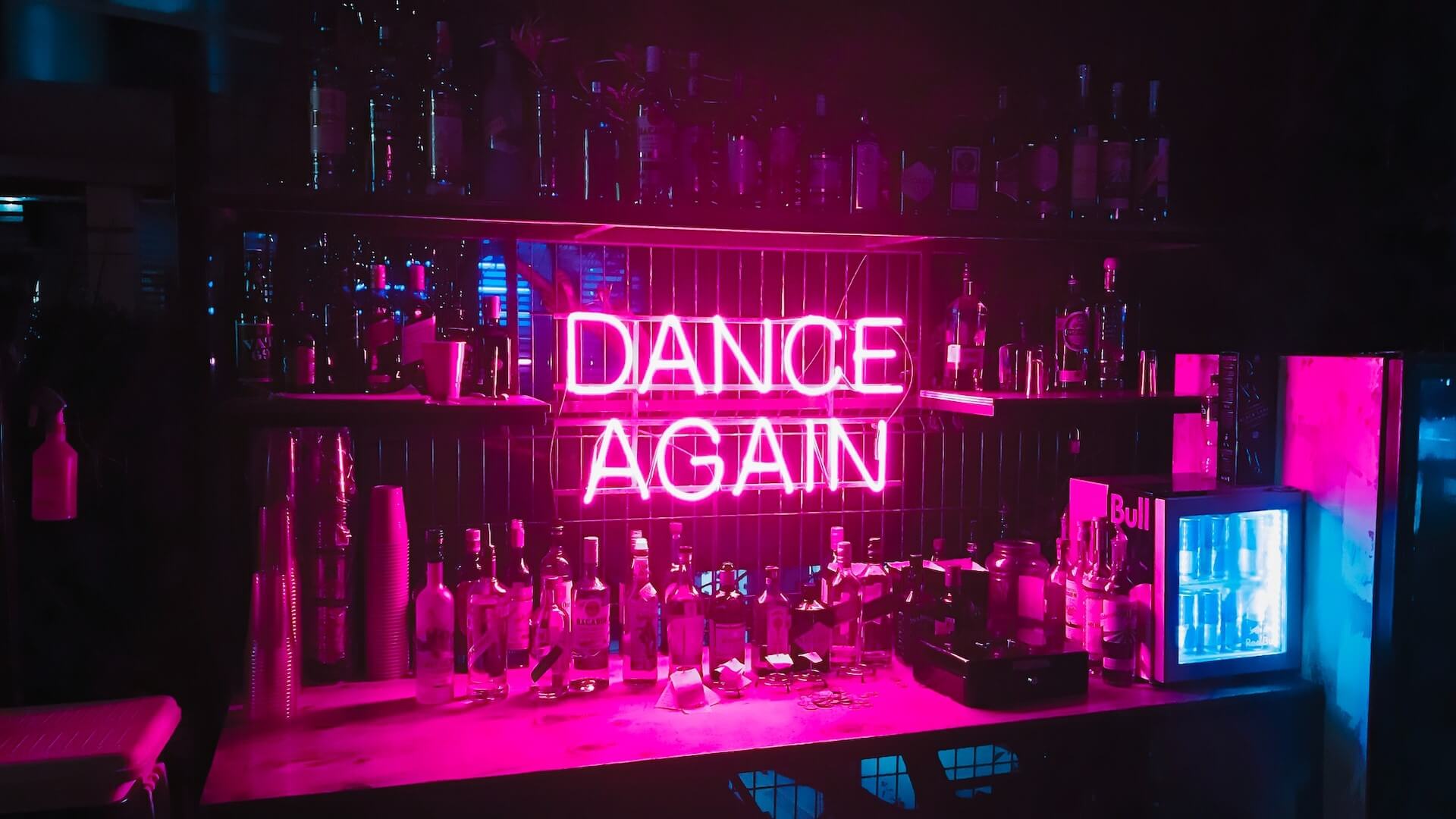 Un bar orné d'étagères contenant des boissons alcoolisées variées et d'une enseigne en néon indiquant 'Dance Again'